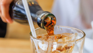 Colaflasche wird in ein Glas mit Strohhalm und Eiswürfeln gegossen