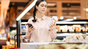 Frau mit Smartphone begutachtet kritisch eine Packung Frau mit Smartphone begutachtet kritisch eine Packung